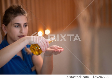 A professional massage therapist pours aromatic oil into their hands, preparing for a relaxing and rejuvenating massage session in a serene spa setting. A professional massage therapist pours aromatic oil into their hands, preparing for a relaxing and rejuvenating massage session in a serene spa setting. 125287200