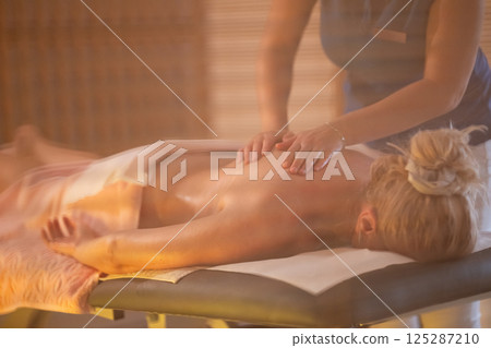 Relaxing Back Massage in a Spa Setting. A professional massage therapist in a blue uniform provides a relaxing back massage to a client lying on a massage table. Relaxing Back Massage in a Spa Setting. A professional massage therapist in a blue uniform provides a relaxing back massage to a client lying on a massage table. 125287210