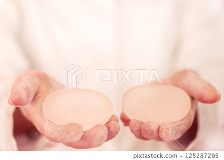 Close-up of a woman hands in transparent gloves holding silicone round breast implants. Breast modern endoprostheses are placed on the palms of the female surgeon. 125287295