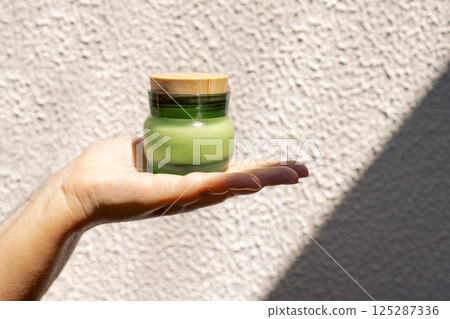 Close-up of a female hand with a jar of green frosted glass moisturizer with a wooden lid. A young woman holds a cosmetic product cream. 125287336