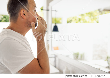 A young man in a white T-shirt puts cream on his cheek in the bathroom. A male looks at himself in the mirror. The man is focused on his skin care routine. 125287340