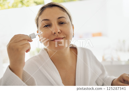 Close-up of face of young woman applying serum to her skin. Natural lady in white robe with pipette with moisturizing nourishing essence. 125287341
