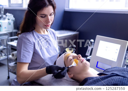 A young woman does a cosmetic procedure at a beauty center or clinic. The cosmetologist applies a rejuvenating orange mask to the client face. 125287350