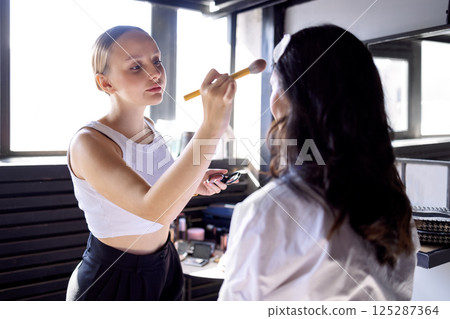 A charming blonde makeup artist applies makeup to her attractive client A charming blonde makeup artist applies makeup to her attractive client 125287364
