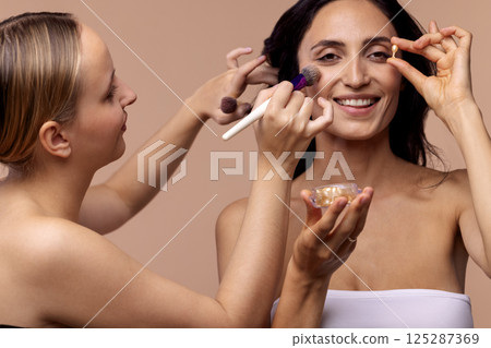 Charming blonde makeup artist applies makeup to her attractive client. Brunette woman is smiling and holding capsules with moisturizing facial serum. Charming blonde makeup artist applies makeup to her attractive client. Brunette woman is smiling and holding capsules with moisturizing facial serum. 125287369