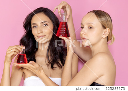 Studio beauty portrait of two beautiful young women with perfect hair and skin. Charming models with glass flasks with red liquid on a pink background. Studio beauty portrait of two beautiful young women with perfect hair and skin. Charming models with glass flasks with red liquid on a pink background. 125287373