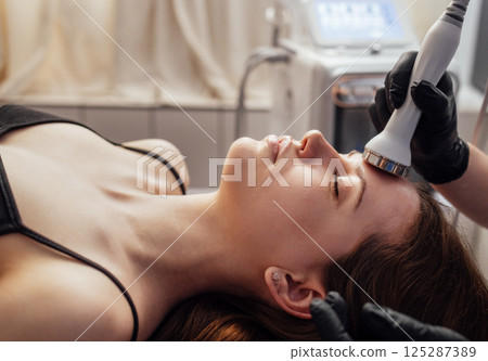 Cream for ultrasonic facial massage is applied to face of young woman using special nozzle in beauty salon 125287389