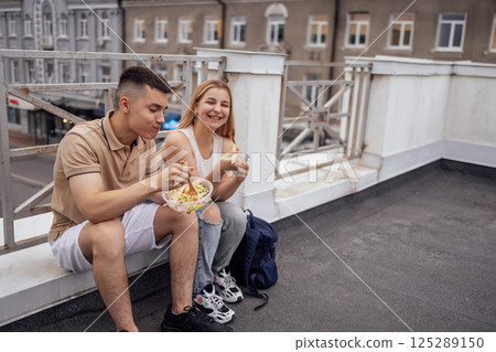 Two teenagers are sitting on the parapet on the roof of the house and eating Asian food 125289150