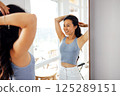 A young attractive Asian woman in casual clothes combs in front of the mirror at home. A charming female Korean smiles and does her hair in the living room. 125289151