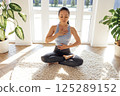 Young attractive Asian woman is sitting in lotus position on carpet in living room. Charming girl in fitness suit meditates and monitors her breathing during yoga class. 125289152