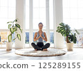Young beautiful Asian woman has closed her eyes and is sitting in lotus position on round carpet in living room. Attractive girl in fitness suit meditates during yoga class. 125289155
