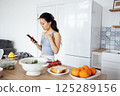 Young attractive Asian woman uses her phone and searches for recipe for dish on Internet. Charming female Korean cooks food in kitchen at home. 125289156
