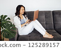 Attractive Asian freelancer is sitting on the sofa in the living room and working on a laptop. Smiling Korean woman is chatting with friends. 125289157