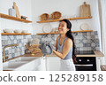 Smiling attractive Asian woman in casual clothes wipes her hands with a towel. A charming Korean woman cleans up the kitchen and laughs. 125289158