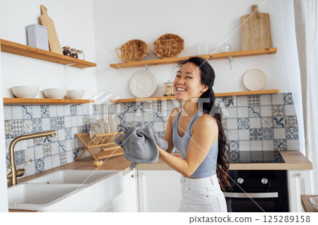 Smiling attractive Asian woman in casual clothes wipes her hands with a towel. A charming Korean woman cleans up the kitchen and laughs. Smiling attractive Asian woman in casual clothes wipes her hands with a towel. A charming Korean woman cleans up the kitchen and laughs. 125289158