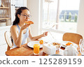 Young attractive Asian woman in glasses and casual clothes is sitting at table and eating sandwich. Charming girl is having breakfast in kitchen at home. 125289159