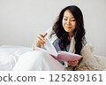 An attractive young Asian woman is lying in bed on pillows and reading a book. A enchanting female Korean is resting on the sofa in the bedroom. 125289161