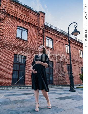 Elegant pregnant woman poses outdoors in a stylish black dress near historic brick buildings at dusk Elegant pregnant woman poses outdoors in a stylish black dress near historic brick buildings at dusk 125289432