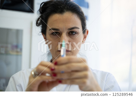 A focused nurse in a white lab coat carefully prepares a syringe for an injection in a professional healthcare environment. 125289546