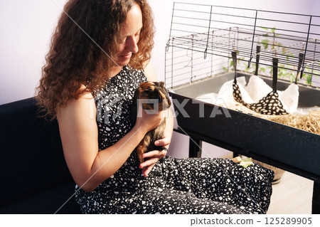Woman holds a guinea pig while sitting near its cage in a cozy indoor setting during daylight hours Woman holds a guinea pig while sitting near its cage in a cozy indoor setting during daylight hours 125289905