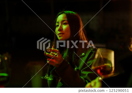 Woman enjoying a drink during a lively gathering in a dimly lit setting with vibrant green lights at night Woman enjoying a drink during a lively gathering in a dimly lit setting with vibrant green lights at night 125290051