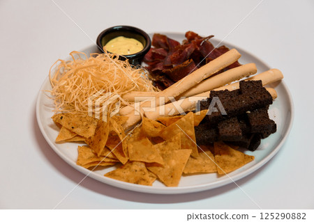 Assorted snacks plate with tortilla chips, breadsticks, jerky, angel hair pasta and yellow sauce in black bowl on white background 125290882