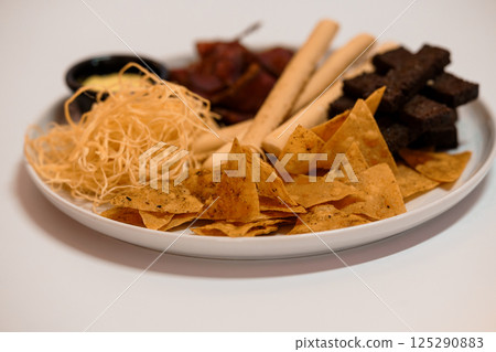 Assorted snack plate with tortilla chips, beef jerky, breadsticks, string cheese and dark chocolate cubes on a white background 125290883
