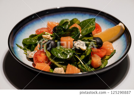 Fresh salad with salmon, spinach, tomatoes, olives and lemon served in blue ceramic bowl on white surface 125290887