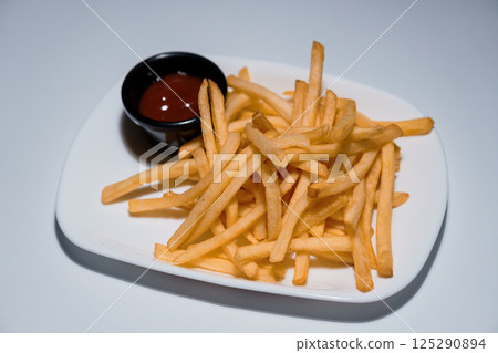 Golden crispy french fries served on a white rectangular plate with a small black bowl of ketchup sauce, isolated on light background 125290894