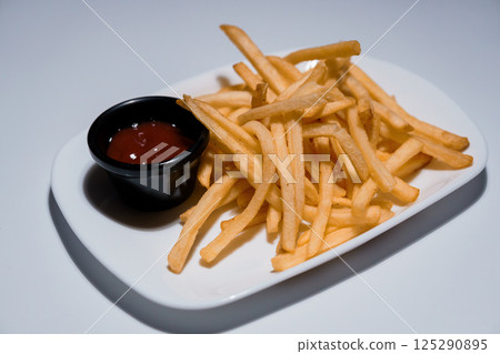 Golden crispy french fries served on a white rectangular plate with a small black bowl of ketchup dipping sauce, studio shot 125290895