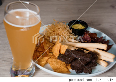 Glass of beer with a plate of snacks including tortilla chips, jerky, breadsticks, and a small bowl of mustard on a wooden surface Glass of beer with a plate of snacks including tortilla chips, jerky, breadsticks, and a small bowl of mustard on a wooden surface 125290916