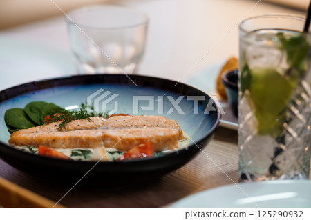 Delicious salmon fillet served on a blue plate with spinach, tomatoes and dill, alongside a refreshing drink in a restaurant setting 125290932