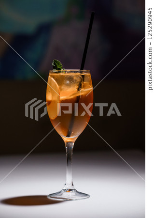 Refreshing cocktail in a wine glass with orange slice, ice cubes, mint garnish, and black straw on white surface with dark blurred background Refreshing cocktail in a wine glass with orange slice, ice cubes, mint garnish, and black straw on white surface with dark blurred background 125290945