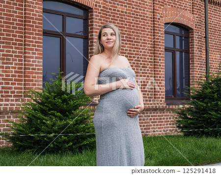 Pregnant woman cradles her baby bump while standing outdoors by a brick building Pregnant woman cradles her baby bump while standing outdoors by a brick building 125291418