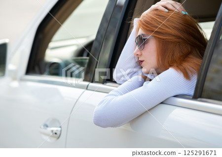 Young woman with red hair and sun glasses travelling by car. Passenger looking out of the rear window of a taxi in a city. Young woman with red hair and sun glasses travelling by car. Passenger looking out of the rear window of a taxi in a city. 125291671