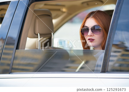 Young woman with red hair and sun glasses travelling by car. Passenger looking out of the rear window of a taxi in a city. Young woman with red hair and sun glasses travelling by car. Passenger looking out of the rear window of a taxi in a city. 125291673