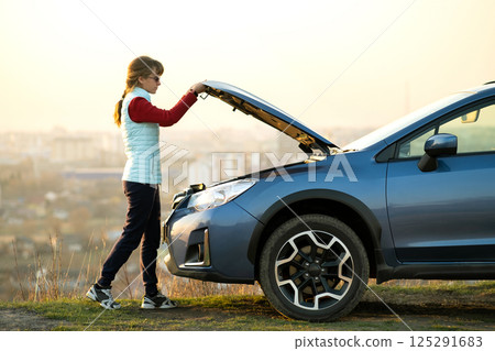 Young woman opening bonnet of broken down car having trouble with her vehicle. Female driver standing near auto with popped up hood. Young woman opening bonnet of broken down car having trouble with her vehicle. Female driver standing near auto with popped up hood. 125291683