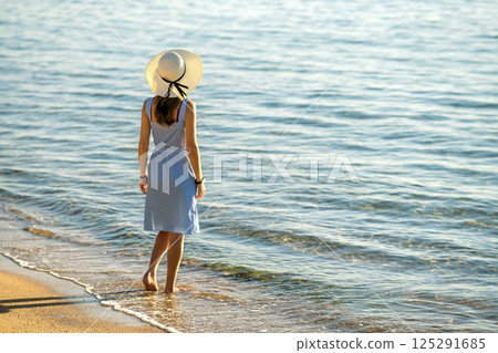 Young woman in straw hat and a dress standing alone on empty sand beach at sea shore. Lonely tourist girl looking at horizon over calm ocean surface on vacation trip. Young woman in straw hat and a dress standing alone on empty sand beach at sea shore. Lonely tourist girl looking at horizon over calm ocean surface on vacation trip. 125291685