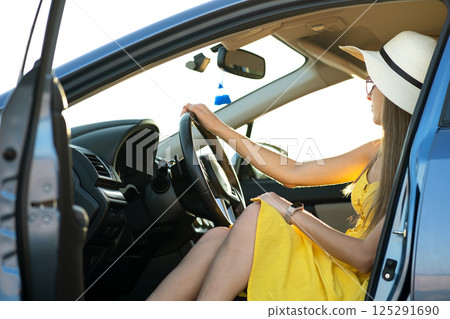 Young woman driver wearing yellow dress and straw hat sitting behind steering wheel driving a car. Summer vacation and travel concept. 125291690