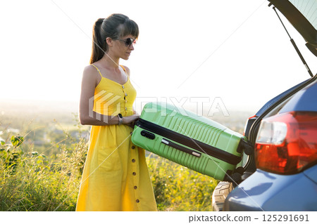 Young woman driver taking green suitcase out from car trunk. Travel and vacations concept. 125291691