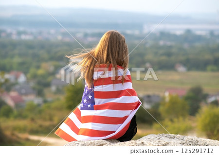 Young pretty american woman with long hair holding waving on wind USA flag on her sholders resting outdoors enjoying warm summer day. 125291712