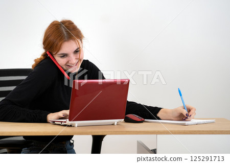 Young office worker woman talking on a cell phone sitting behind working desk with laptop computer and notebook. 125291713