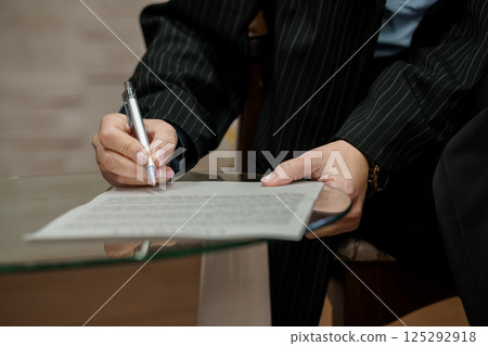 Person Signing Document on Glass Table with Pen in Business Attire Person Signing Document on Glass Table with Pen in Business Attire 125292918