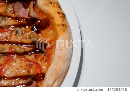 Close-up of a delicious pizza with barbecue sauce, chicken strips, tomatoes, mushrooms, and ham on a white plate 125293016