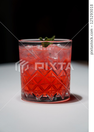 Refreshing red cocktail in a faceted glass with ice and mint garnish on white surface, dark background 125293018