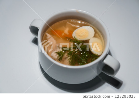 Chicken noodle soup in a white bowl with two halves of boiled egg and dill, served on light background 125293023