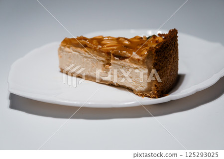 Slice of delicious caramel cheesecake with peanut topping on a white plate against a plain background, close-up view 125293025