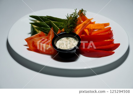 Colorful vegetable platter with fresh sliced tomatoes, cucumbers, carrots, red peppers and creamy dip served on a white plate 125294508