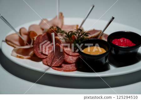 Appetizing assortment of sliced cured meats, including salami and ham, served with mustard and berry sauce on a white plate 125294510