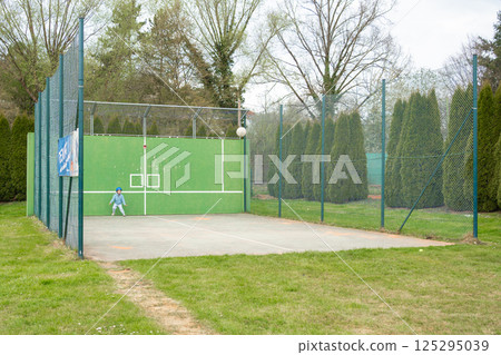 Child in sports outfit plays with a ball at a green-walled court. Natural setting, solo action, and clear space perfect for campaigns on childhood development, sports safety, or active lifestyle. 125295039